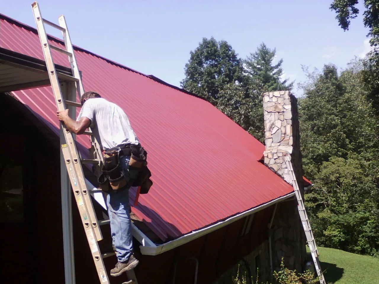 Roofer on a ladder beside a red metal roof and stone chimney — Soffit & Fascia Repair by Roofing Brothers in Rich Square